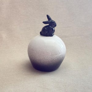 May include: A ceramic decorative piece featuring a black bunny figurine atop a white and gray speckled sphere. The sphere transitions from white to a dark gray at the base, creating a gradient effect. The bunny is detailed with visible ears and a seated pose.