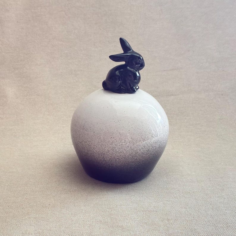 May include: A ceramic decorative piece featuring a black bunny figurine atop a white and gray speckled sphere. The sphere transitions from white to a dark gray at the base, creating a gradient effect. The bunny is detailed with visible ears and a seated pose.