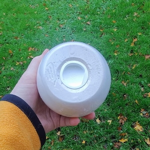 May include: A light gray, spherical object with a circular opening. The object is held in a hand against a backdrop of green grass and fallen leaves. The object appears to be made of ceramic or a similar material.