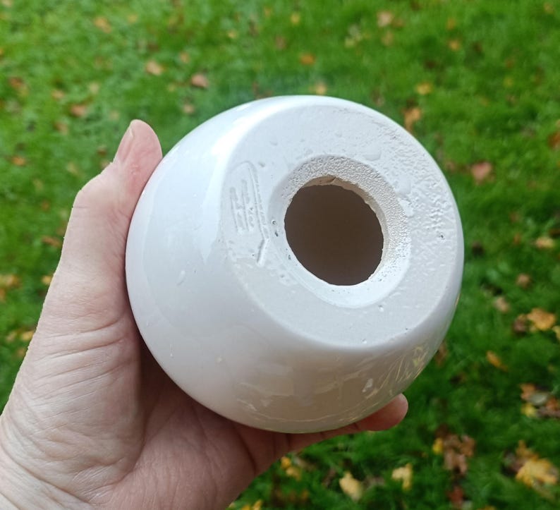 May include: A white, spherical ceramic object with a large central opening. The object has a glossy finish and appears to be a decorative item or a part of a larger piece. The base has a rough, unglazed texture.