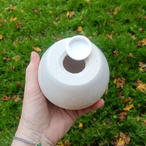 May include: A white, spherical ceramic vase with a circular opening and a small, attached lid. The vase is held in a hand against a backdrop of green grass and fallen leaves. The vase has a smooth, matte finish.