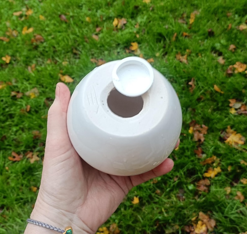 May include: A white, spherical ceramic vase with a circular opening and a small, attached lid. The vase is held in a hand against a backdrop of green grass and fallen leaves. The vase has a smooth, matte finish.