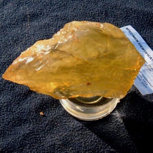 May include: A translucent, amber-colored specimen of desert glass, resting on a clear glass container. The desert glass has a rough, irregular shape and is partially illuminated by sunlight. A small tag with text is visible.