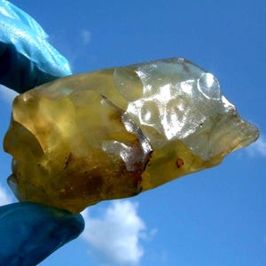 Pode incluir: Uma gema translúcida de cor âmbar com uma forma irregular e áspera. A pedra tem uma superfície ligeiramente brilhante e parece ser segurada por uma mão enluvada contra um fundo de céu azul brilhante com nuvens dispersas. A gema tem algumas inclusões escuras.