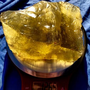 May include: A large, translucent, yellow-green mineral specimen is displayed on a digital scale. The scale reads 1265, likely indicating the weight in grams. The mineral has a rough, uneven surface and a crystalline appearance.