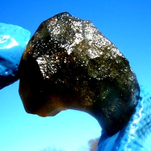 Op de afbeelding: Een donkere, onregelmatig gevormde steen met een ruw, getextureerd oppervlak. De steen heeft een donkere, bijna zwarte kleur met sprankelende, reflecterende insluitsels. De achtergrond is helderblauw.