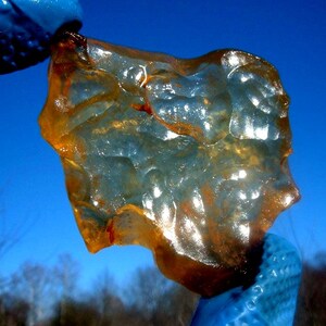 May include: A translucent, irregular piece of amber-coloured resin with a glossy, undulating surface texture. The material displays varying shades of golden yellow and deep orange, with light passing through to reveal internal clarity.