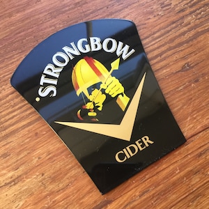 May include: Black and gold Strongbow Cider sign featuring a graphic of a person wearing a helmet and holding a bow and arrow.