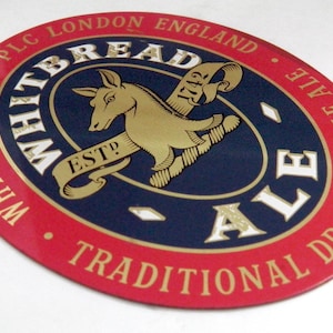 May include: An oval-shaped Whitbread Ale beer coaster featuring a red border with gold text reading Whitbread & Co. PLC London England and Traditional Draught Pale Ale. The center is dark blue with a gold horse graphic and the text Est 1742.