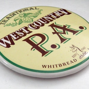 May include: A round, cream-colored ceramic beer tap handle featuring the text "TRADITIONAL WEST COUNTRY P.A." in dark brown and green. It includes a green hop illustration, a small horse graphic with "ESTD 1742", and the word "WHITBREAD & COMPANY".