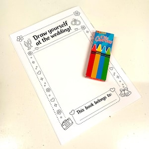 May include: A white paper activity sheet with a black outline border featuring wedding-themed icons like rings, flowers, and bells. The text reads Draw yourself at the wedding and This book belongs to. A small box of four wax crayons is placed on top.