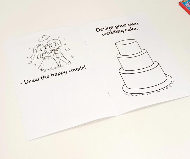 May include: An open activity book with white pages featuring black line art. The left page shows a cartoon couple with the text Draw the happy couple! The right page displays a three-tiered wedding cake with the text Design your own wedding cake.
