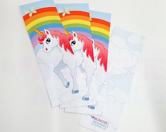 Könnte beinhalten: Drei weiße Einhorn-Lesezeichen mit rosafarbenen Mähnen und Hörnern. Jedes Lesezeichen zeigt einen Regenbogen und Wolken auf blauem Hintergrund. Die Lesezeichen sind mit dem Text "Munchie Moodskids" und einer Website-Adresse bedruckt.