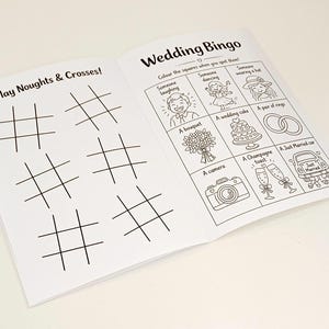 May include: An open activity booklet with black and white line art. The left page features six grids for Play Noughts & Crosses. The right page is titled Wedding Bingo and contains a grid with illustrations of a laughing person, dancing person, hat, bouquet, cake, rings, camera, champagne, and a car.
