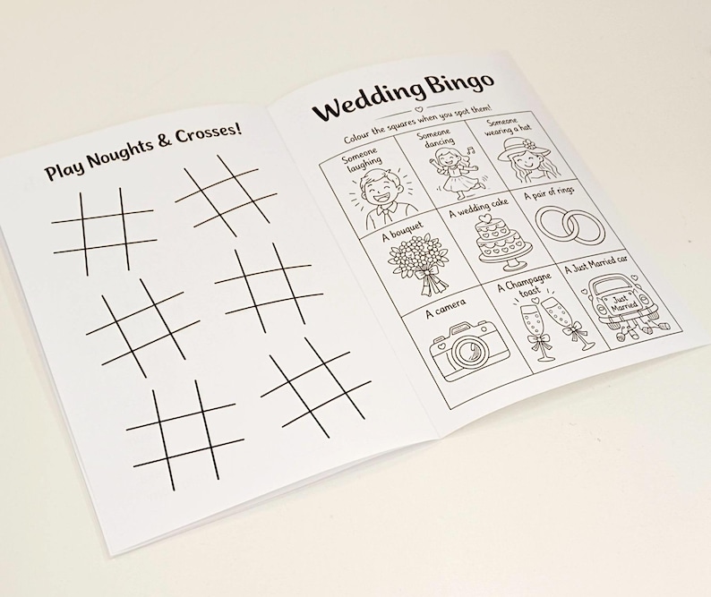 May include: An open activity booklet with black and white line art. The left page features six grids for Play Noughts & Crosses. The right page is titled Wedding Bingo and contains a grid with illustrations of a laughing person, dancing person, hat, bouquet, cake, rings, camera, champagne, and a car.