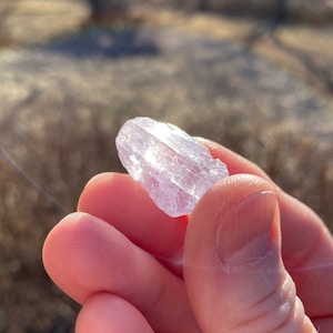 May include: A translucent, light purple crystal with a pointed end. The crystal is held in a hand.
