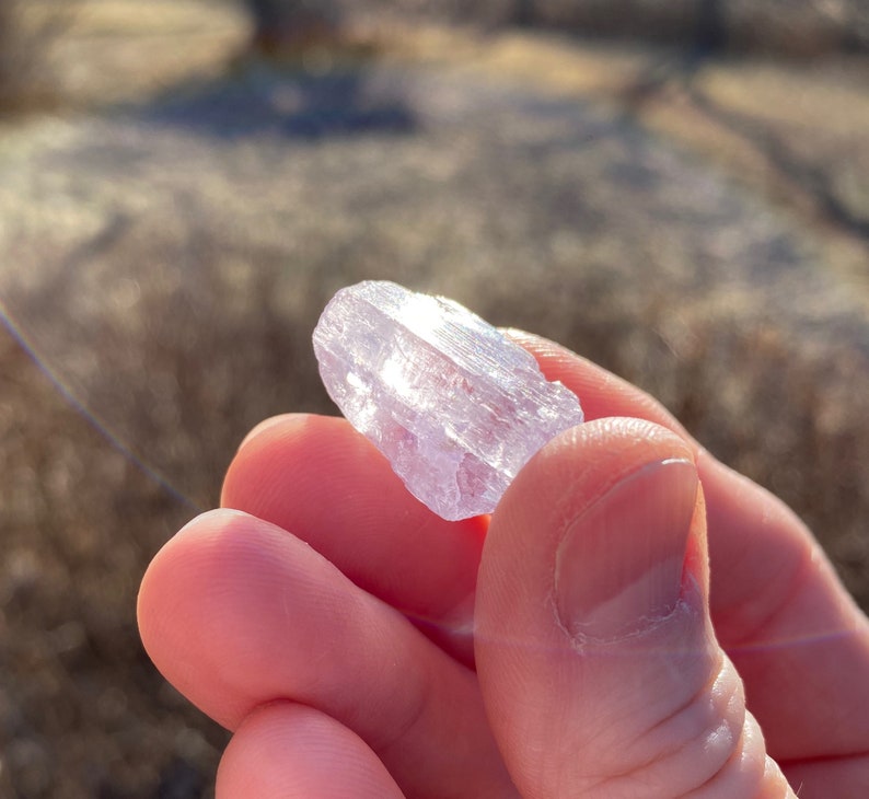May include: A translucent, light purple crystal with a pointed end. The crystal is held in a hand.