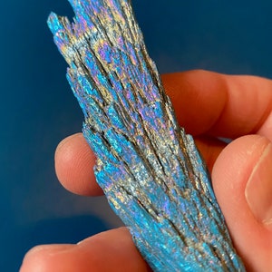 May include: An iridescent blue and purple crystal with a layered, striated texture. The crystal is held in a hand.