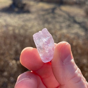 May include: A small, translucent, light pink crystal with a rough, natural texture. The crystal is held in a hand, with the background out of focus.