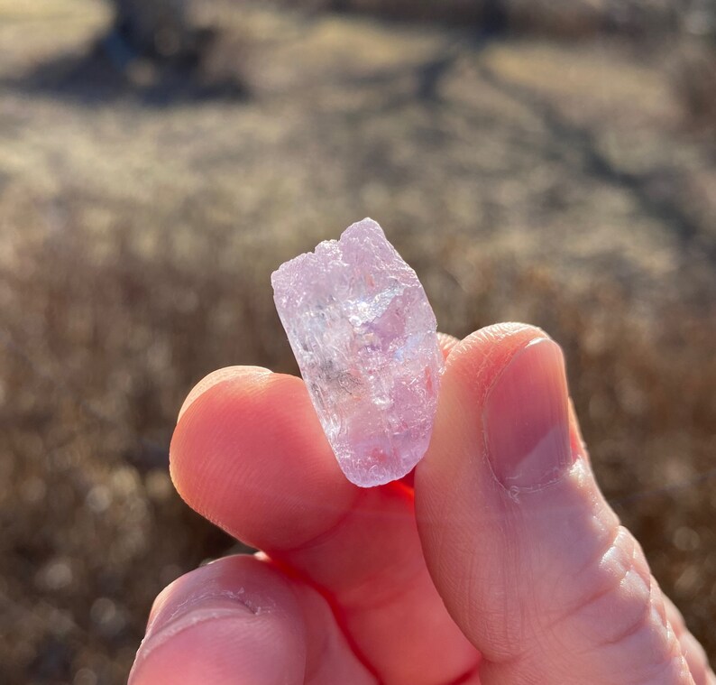 May include: A small, translucent, light pink crystal with a rough, natural texture. The crystal is held in a hand, with the background out of focus.