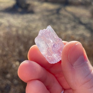 May include: A translucent, pale purple crystal with a striated texture. The crystal is held in a hand.