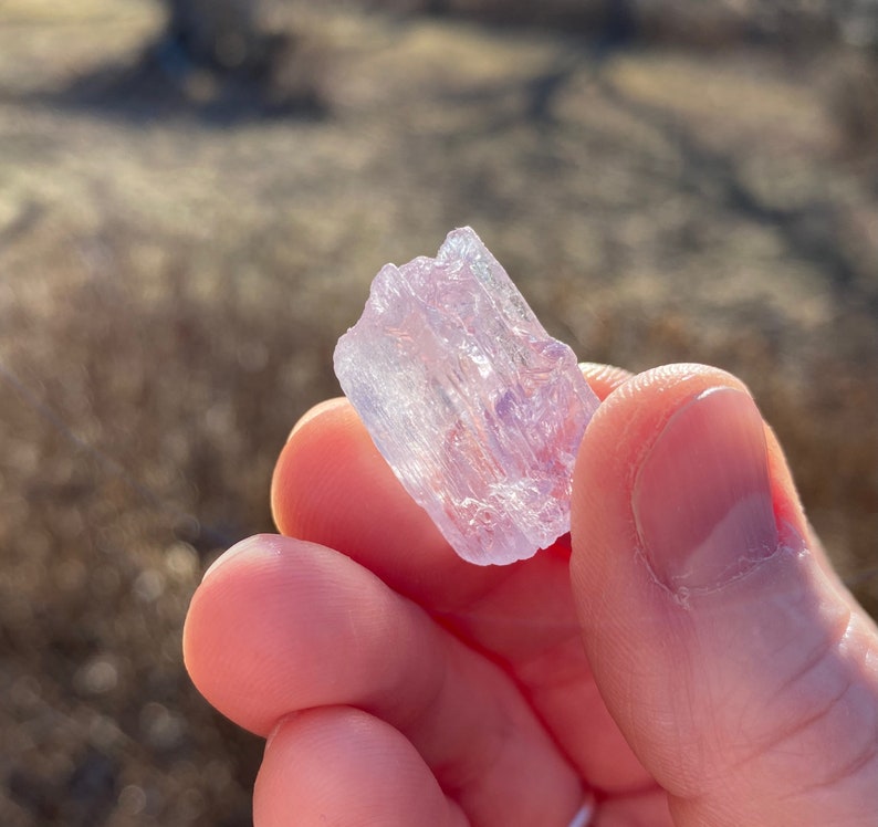 May include: A translucent, pale purple crystal with a striated texture. The crystal is held in a hand.