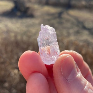May include: A clear, translucent crystal with a light purple hue. The crystal is shaped like a prism and has many facets. It is being held in a hand.