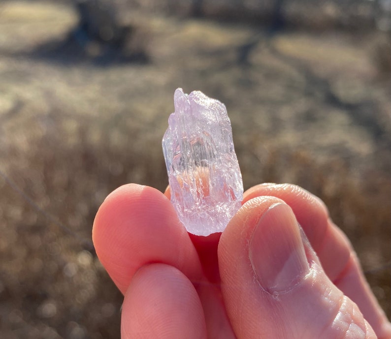 May include: A clear, translucent crystal with a light purple hue. The crystal is shaped like a prism and has many facets. It is being held in a hand.