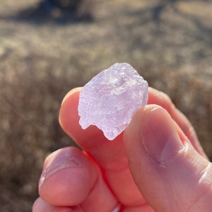 May include: A close-up of a pale purple crystal with a rough, irregular surface. The crystal is being held by a hand.