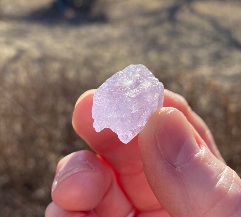May include: A close-up of a pale purple crystal with a rough, irregular surface. The crystal is being held by a hand.