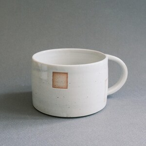 Könnte beinhalten: Eine weiße Keramiktasse mit rundem Korpus und Henkel. Die Tasse hat ein kleines, quadratisches, braunes Design an der Seite. Die Tasse steht vor einem grauen Hintergrund, der ihr schlichtes, minimalistisches Design hervorhebt.