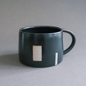 Könnte beinhalten: Eine dunkelblaue Keramiktasse mit rundem Körper und gebogenem Henkel. Die Tasse hat einen rechteckigen beigen Akzent und einen vertikalen weißen Streifen. Das Innere der Tasse ist schwarz.