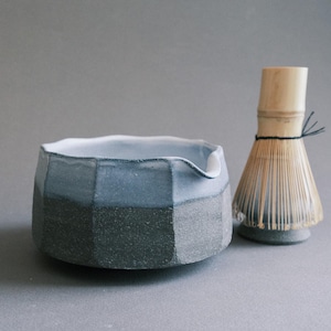 Peut inclure: Un bol à matcha en céramique avec un design à facettes, présentant des tons bleus et gris. À côté, un fouet à matcha en bambou, outil traditionnel pour préparer le thé matcha. Le bol a un bec verseur.