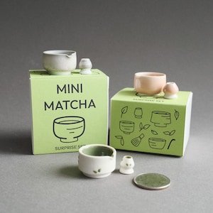 May include: Two light green cardboard boxes labeled MINI MATCHA SURPRISE SET with line art illustrations. Each box holds a miniature ceramic bowl and whisk. A third bowl and whisk set sits in front with a small round metal coin for scale.