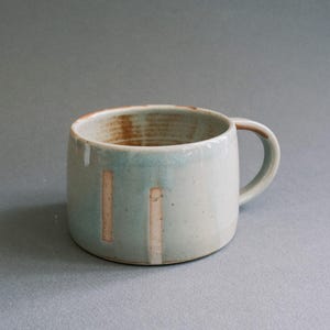 May include: A handmade ceramic mug with a light blue glaze and two vertical beige stripes. The interior has a brown, textured appearance. The mug has a rounded shape and a small handle. This mug is perfect for coffee or tea.