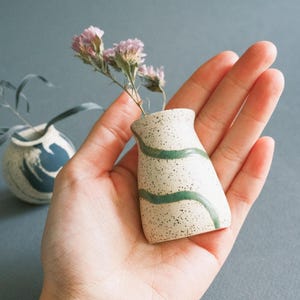 May include: A small, speckled ceramic vase with green wavy lines, held in a hand. The vase contains dried pink flowers. Another small vase with blue and white design is in the background. The items are on a gray surface.