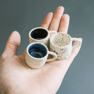 May include: Three small ceramic mugs with handles, each with a speckled, off-white exterior. The interior of each mug is a different color: dark blue, white, and navy blue. The mugs are held in a hand against a gray background.