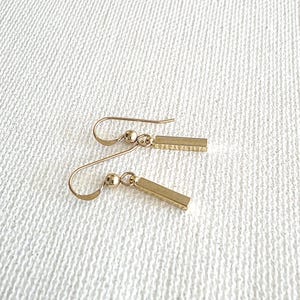 May include: A pair of gold-toned drop earrings featuring a small spherical bead attached to a slender, rectangular bar pendant. The earrings are suspended from curved ear wires, showcasing a polished metallic finish and a simple, linear design.