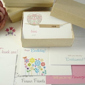 May include: A gold-toned bar necklace engraved with the date 7-11-15, presented in a tan cardboard gift box. The box includes a card with a heart illustration and the text I love you, surrounded by various greeting cards with floral designs.
