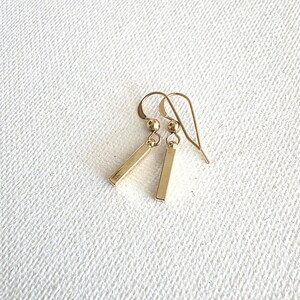 May include: A pair of gold-toned drop earrings featuring a slender, rectangular bar pendant suspended from a small spherical bead and a curved ear wire hook. The earrings have a polished, metallic finish and a simple, linear design.