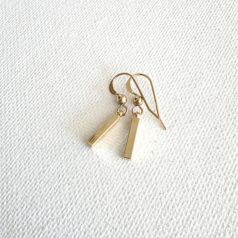 May include: A pair of gold-toned drop earrings featuring a slender, rectangular bar pendant suspended from a small spherical bead and a curved ear wire hook. The earrings have a polished, metallic finish and a simple, linear design.