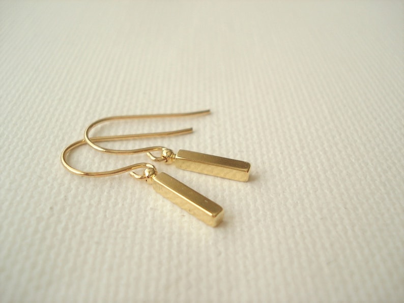 May include: A pair of gold-toned drop earrings featuring small, smooth, rectangular bar pendants suspended from curved ear wires. The metal has a polished, reflective finish, and the design consists of two identical pieces linked by a small jump ring.