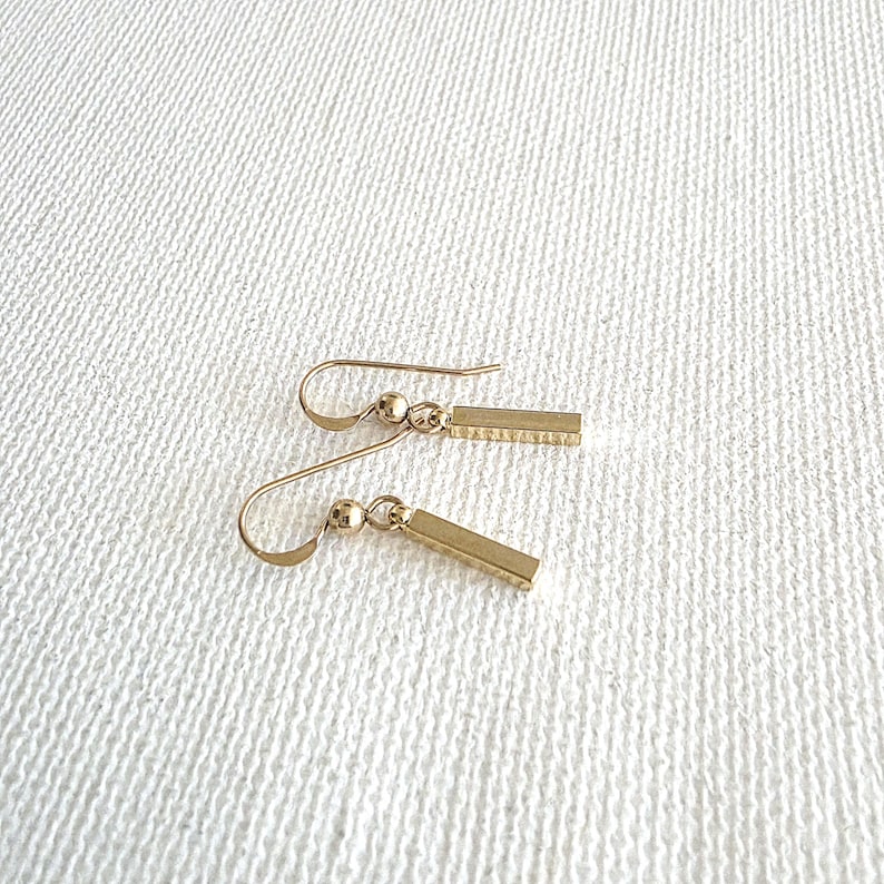 May include: A pair of gold-toned drop earrings featuring a small spherical bead attached to a slender, rectangular bar pendant. The earrings are suspended from curved ear wires, showcasing a polished metallic finish and a simple, linear design.