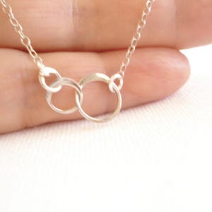Peut inclure: Un délicat collier en argent avec deux cercles entrelacés. Le collier a une fine chaîne et est présenté sur un fond doux et neutre. Les cercles sont reliés, symbolisant l'unité ou la connexion.
