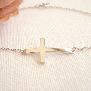 May include: A delicate silver bracelet featuring a sideways cross pendant. The cross is gold-toned and attached to a fine chain. The bracelet is displayed on a white surface, with a soft pink flower in the background.