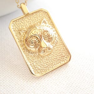 Może przedstawiać: Złoty prostokątny naszyjnik z wisiorkiem z wizerunkiem szczegółowej twarzy tygrysa. Wisiorek ma teksturowane tło i prostokątny kształt. Naszyjnik to biżuteria.