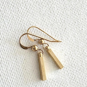 May include: A pair of gold-colored drop earrings. Each earring features a small, round bead connected to a rectangular bar pendant. The earrings have a simple, elegant design with a hook closure.