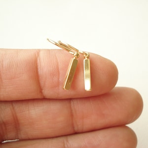 May include: A pair of gold-toned drop earrings featuring slender, rectangular bar pendants suspended from simple curved ear wires. The metallic finish is smooth and reflective, showcasing a clean, geometric design with a polished surface.