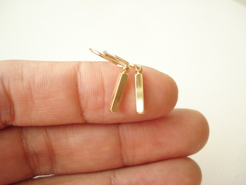 May include: A pair of gold-toned drop earrings featuring slender, rectangular bar pendants suspended from simple curved ear wires. The metallic finish is smooth and reflective, showcasing a clean, geometric design with a polished surface.