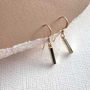 May include: A pair of gold-toned drop earrings featuring a slender, rectangular bar pendant suspended from a curved wire hook. The metal has a polished finish, and the design consists of a simple vertical bar attached to a small jump ring.